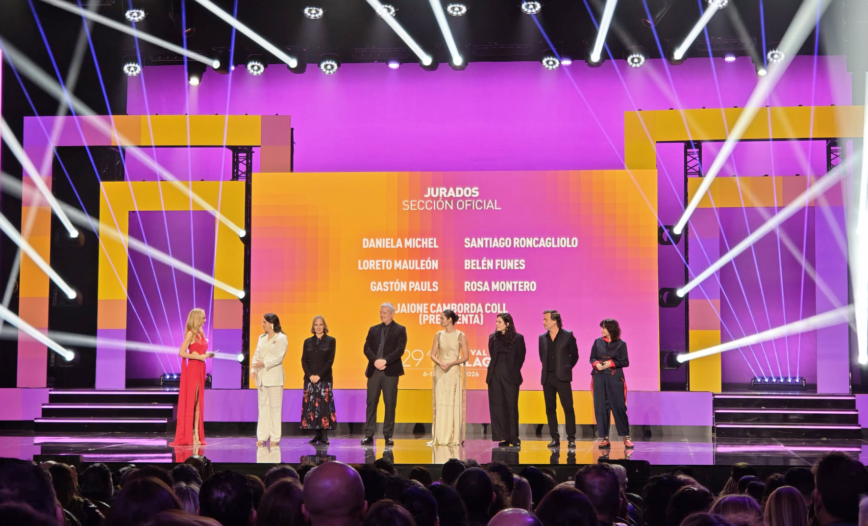 Sobre un escenario iluminado con luces blancas y pantallas, están la actriz española Kira Miró, la Fundadora y directora general del Festival Internacional de Cine de MoreliaDaniela Michel; el escritor y guionista Santiago Roncagliolo; la actriz Loreto Mauleón; la directora Belén Funes; el actor y director Gastón Pauls, y la escritora Rosa Montero.